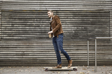 Smiling man with coffee to go on skateboard in front of wooden wall - FMKF03160