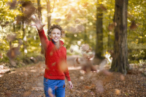 Lächelnder Junge wirft Blätter in die Luft im herbstlichen Wald, lizenzfreies Stockfoto