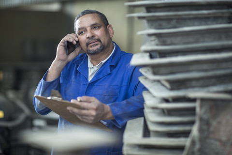 Mann mit Klemmbrett beim Telefonieren in einer Fabrik, lizenzfreies Stockfoto