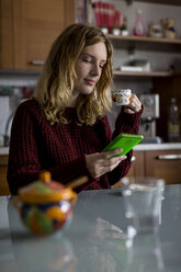 Frau mit Tasse Kaffee sitzt in der Küche und schaut auf ihr Smartphone - MAUF00853