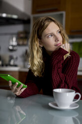 Nachdenkliche Frau sitzt in der Küche mit Smartphone und Tasse Tee - MAUF00858