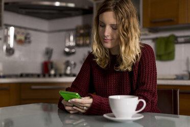 Frau sitzt in der Küche mit einer Tasse Tee und benutzt ein Mobiltelefon - MAUF00859