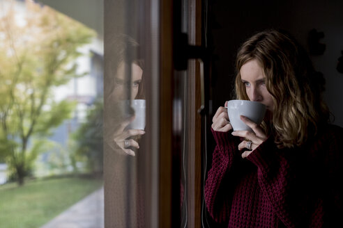 Nachdenkliche Frau trinkt eine Tasse Tee und schaut durch ein Fenster - MAUF00860