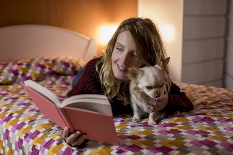 Lächelnde Frau auf dem Bett liegend mit ihrem Hund, der ein Buch liest, lizenzfreies Stockfoto
