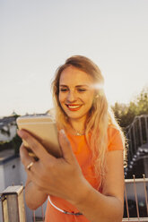 Porträt einer lächelnden blonden Frau, die ein Selfie auf einer Dachterrasse mit Gegenlicht macht - AIF00417
