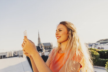 Österreich, Wien, lächelnde blonde Frau macht Selfie auf Dachterrasse mit Stephansdom im Hintergrund - AIF00418
