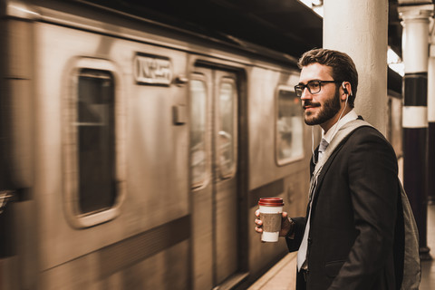 Junger Geschäftsmann wartet am Bahnsteig einer U-Bahn-Station und hält einen Einwegbecher, lizenzfreies Stockfoto