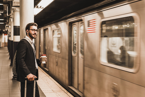 Junger Geschäftsmann wartet am Bahnsteig einer U-Bahn-Station und hält einen Einwegbecher, lizenzfreies Stockfoto