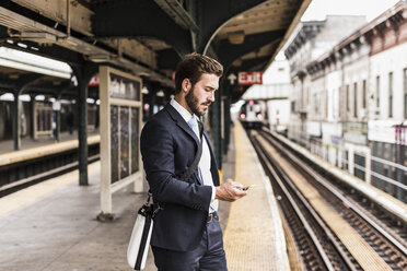 Junger Geschäftsmann, der am Bahnsteig einer U-Bahn-Station wartet und ein Smartphone benutzt - UUF09021