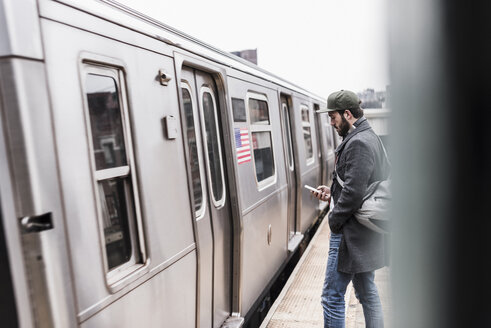 Junger Mann, der am Bahnsteig auf die U-Bahn wartet und sein Smartphone benutzt - UUF09036
