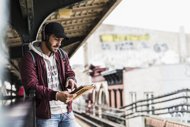 Junger Mann, der auf die U-Bahn wartet, Kopfhörer trägt und ein digitales Tablet benutzt - UUF09045