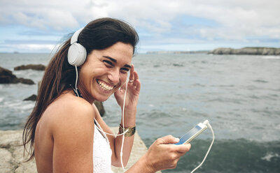 Lachende Frau hört Musik mit Kopfhörern vor dem Meer - DAPF00463