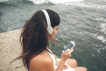 Frau hört Musik mit Kopfhörern und schaut auf ihr Smartphone vor dem Meer - DAPF00464