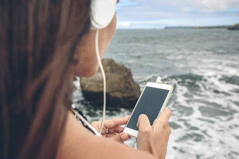 Frau, die ihr Smartphone benutzt, während sie mit Kopfhörern vor dem Meer hört, lizenzfreies Stockfoto