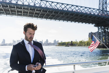USA, New York City, Geschäftsmann auf Fähre auf dem East River, der sein Mobiltelefon überprüft - UUF09053