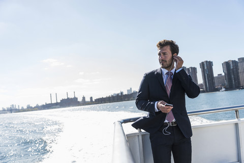 USA, New York City, businessman on ferry on East River with cell phone and earphones stock photo