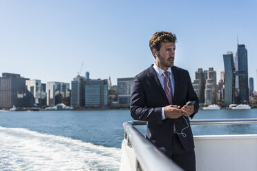 USA, New York City, businessman on ferry on East River with cell phone and earphones - UUF09064