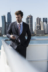 USA, New York City, businessman on ferry on East River with cell phone and earphones - UUF09070