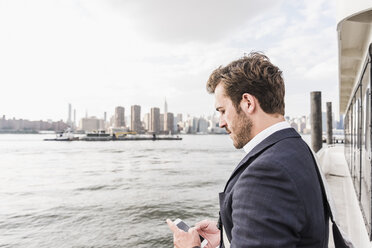 USA, New York City, Geschäftsmann auf Fähre auf dem East River, der sein Mobiltelefon überprüft - UUF09110