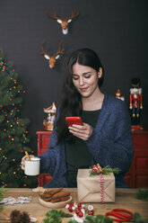 Smiling young woman with cup of coffee lokking at her cell phone at home - RTBF00500