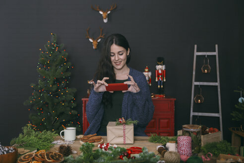 Lächelnde junge Frau, die ein dekoriertes Weihnachtsgeschenk mit ihrem Smartphone fotografiert - RTBF00501