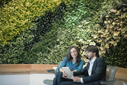 Geschäftsmann und Frau sitzen vor einer grünen Pflanzenwand und benutzen ein digitales Tablet - WESTF21888