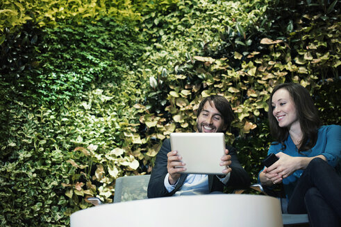Geschäftsmann und Frau sitzen vor einer grünen Pflanzenwand und benutzen ein digitales Tablet - WESTF21891
