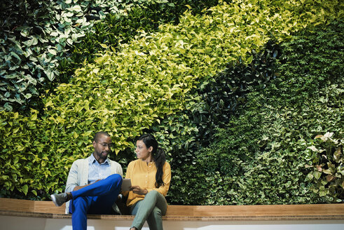 Businessman and woman sitting in front of green plant wall, using digital tablet - WESTF21898