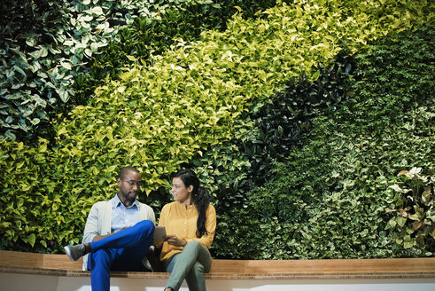 Geschäftsmann und Frau sitzen vor einer grünen Pflanzenwand und benutzen ein digitales Tablet - WESTF21898