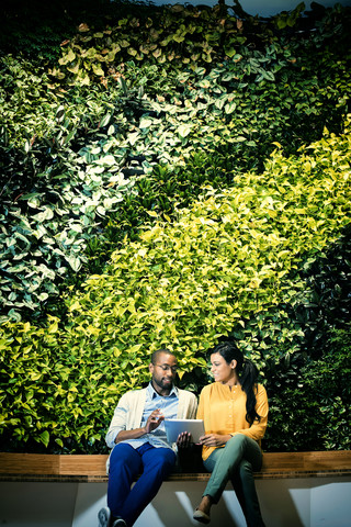 Geschäftsmann und Frau sitzen vor einer grünen Pflanzenwand und benutzen ein digitales Tablet, lizenzfreies Stockfoto