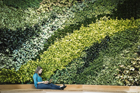 Junge Frau mit Laptop vor einer grünen Pflanzenwand, lizenzfreies Stockfoto