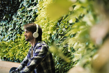 Junger Mann sitzt vor einer grünen Pflanzenwand und trägt Kopfhörer - WESTF21907