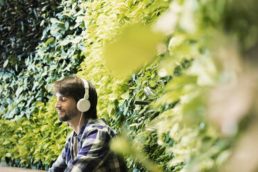 Junger Mann sitzt vor einer grünen Pflanzenwand und trägt Kopfhörer - WESTF21908