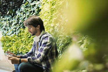 Junger Mann sitzt vor einer grünen Pflanzenwand und benutzt ein digitales Tablet - WESTF21909