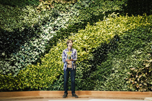 Young man standing in front of green plant wall, holding potted plant - WESTF21912