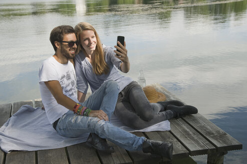 Lächelndes junges Paar sitzt auf einem Steg an einem See und macht ein Selfie - CRF02761