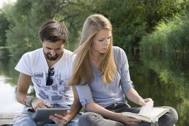 Junges Paar sitzt mit Buch und Tablet auf einem Steg an einem See - CRF02765