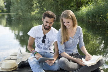 Smiling young couple sitting on jetty at a lake with book and tablet - CRF02766