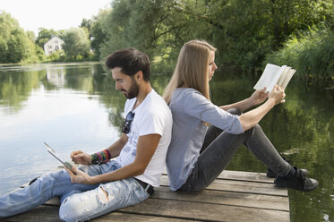 Junges Paar sitzt mit Buch und Tablet auf einem Steg an einem See - CRF02767