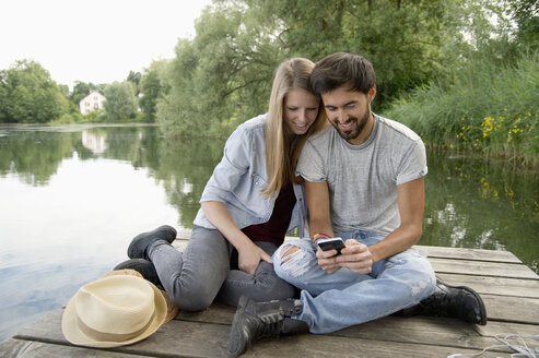 Lächelndes junges Paar sitzt mit Handy auf einem Steg am See - CRF02771