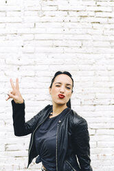 Young woman in front of brick wall making victory sign and pouting - GEMF01190