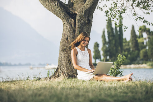 Italien, Gardasee, junge Frau lehnt an einem Baum und benutzt einen Laptop - SBOF00267
