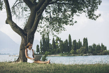 Italy, Lake Garda, young woman leaning against a tree using laptop - SBOF00268