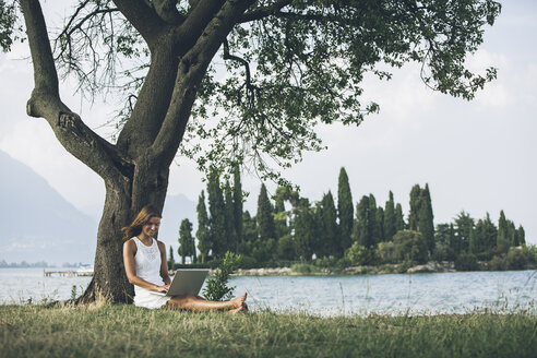 Italien, Gardasee, junge Frau lehnt an einem Baum und benutzt einen Laptop - SBOF00268