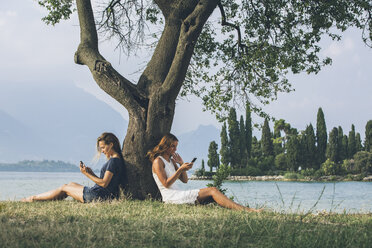 Italien, Gardasee, zwei junge Frauen lehnen an einem Baum und benutzen Handys - SBOF00277