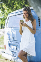 Lächelnde junge Frau lehnt an einem Campingbus und schaut auf ihr Handy - SBOF00293