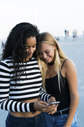 Zwei junge Frauen schauen im Freien auf ihr Handy - KKAF00016