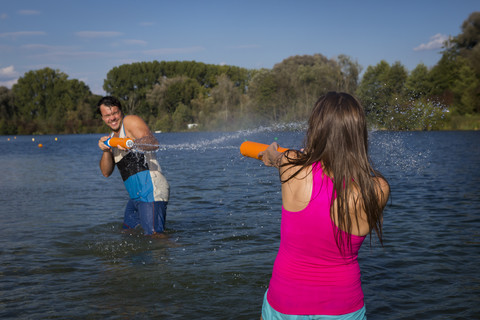 Junges Paar steht am See und spielt mit Wasserpistolen, lizenzfreies Stockfoto