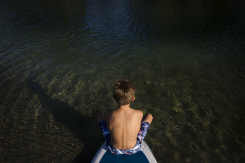Rückenansicht eines Jungen, der auf einem SUP-Board am Seeufer sitzt - JTLF00133