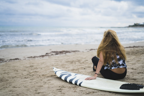 Junge Frau mit Surfbrett am Strand sitzend und auf das Meer blickend, lizenzfreies Stockfoto